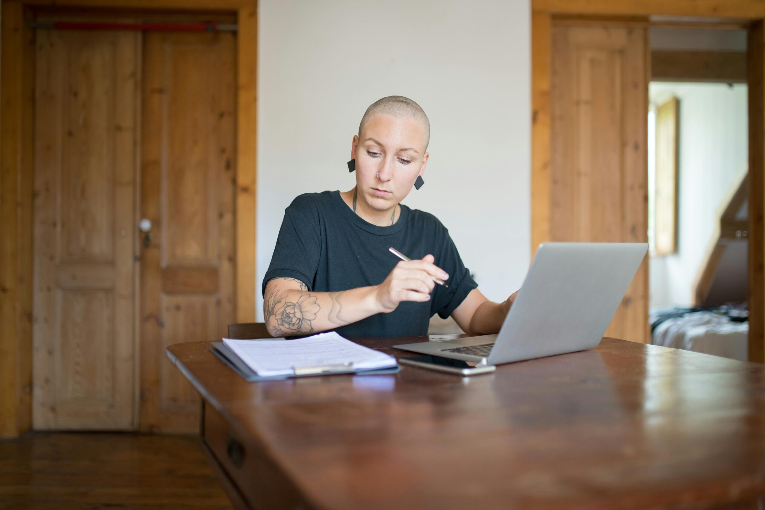 Bald woman with tattoos working from home using a laptop and taking notes. Indoor setting.